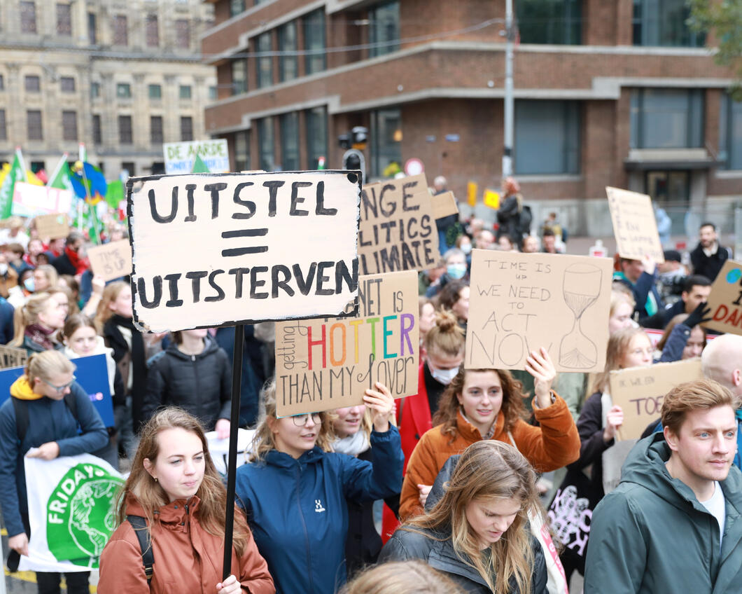 Mensen lopen in de klimaatmars. In het midden houdt iemand een protestbord vast met de tekst Uitstellen is uitsterven. 